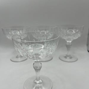 Könnte beinhalten: Vier klare Champagnerkelche aus Glas mit geschliffenem Glasdesign. Die Gläser sind in einer Reihe auf einer weißen Oberfläche angeordnet.