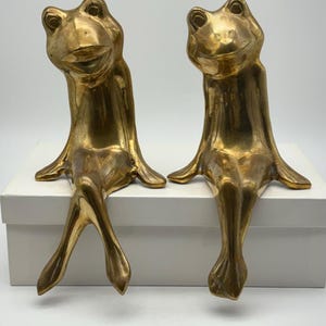 Könnte beinhalten: Zwei goldfarbene Froschfiguren sitzen nebeneinander. Die Frösche haben eine sitzende Haltung mit hängenden Beinen. Die Figuren haben eine glatte, metallische Oberfläche und stehen auf einer weißen Oberfläche.