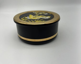 Könnte beinhalten: Eine schwarze Keramik-Schmuckdose mit goldfarbenem Rand und einem goldfarbenen Band um die Mitte. Der Deckel zeigt ein weißes und goldenes Bild eines Vogels in einem Baum.