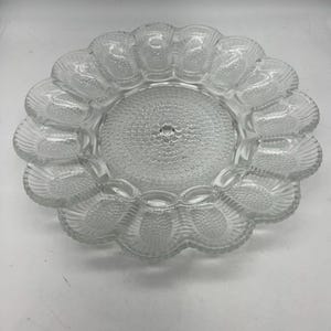 May include: Clear glass egg serving platter with a scalloped edge. The center of the platter has a textured, dotted design. The outer edge has individual egg-shaped compartments.