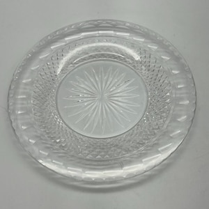 Könnte beinhalten: Eine transparente Glasplatte mit einem Sternmuster in der Mitte und einem Diamantmuster am Rand. Die Platte hat einen leicht erhöhten Rand und ist kreisförmig. Das transparente Glas lässt Licht durchscheinen.