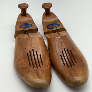 May include: Two wooden shoe trees in a light brown hue. Each has a rounded toe, a heel section, and a knob. The trees feature the text "The T Shoe" on a blue and red label.
