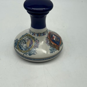 Può includere: Una bottiglia di ceramica bianca con un tappo blu. La bottiglia è decorata con immagini di navi, uno stemma e il testo "St. J. Din La.  La bottiglia è un pezzo decorativo e non è destinata all'uso con liquidi.