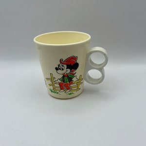Puede incluir: Una taza de plástico blanca con un asa que presenta una imagen de dibujos animados de Mickey Mouse vestido de vaquero. La taza tiene un diseño de valla verde y marrón alrededor de la imagen.