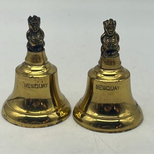 May include: Two gold-colored bells with a small figure on top of each. The bells are inscribed with the word "NEWQUAY".