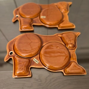 Puede incluir: Dos bandejas de servir de cerámica marrón en forma de vaca con tres hendiduras redondas para salsas o aperitivos. Las bandejas tienen un acabado brillante y están marcadas con "Made in Japan" en la parte inferior.