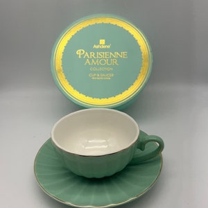 May include: A green teacup and saucer set with a gold rim. The saucer has a scalloped edge. The teacup has a white interior and a green exterior. The set is packaged in a round box with a gold trim and the text "Parisienne Amour Collection Cup & Saucer New Bone China" printed on it.