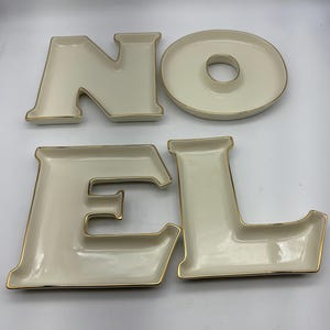 Puede incluir: Un juego de cuatro fuentes de cerámica color crema que deletrean "NOEL" con ribete dorado. La "O" es una fuente circular con un agujero en el centro. Las otras letras son fuentes con forma.