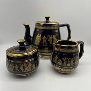 May include: A black and gold tea set comprising a teapot, creamer and sugar bowl. The set features a Greek key design and a scene of dancing figures in gold.