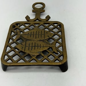 May include: A bronze metal trivet with an anchor-shaped handle. The trivet has a diamond-patterned grid design with two fish silhouettes. It is designed to protect surfaces from hot cookware.