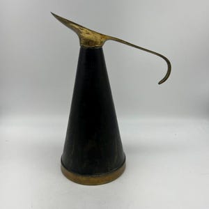 Puede incluir: Una jarra de metal negro y dorado con un pico largo y curvo. La jarra tiene una forma cónica y un borde de latón.
