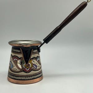 May include: A copper-coloured Turkish coffee pot with a dark wooden handle and a decorative floral pattern in white, red, blue, and yellow. The pot has a wide base and a narrow top, with a silver-coloured rim.