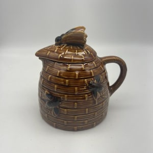 Puede incluir: Un tarro de miel de cerámica marrón con un diseño de colmena y una abeja en la tapa. El tarro tiene una asa y un pico.