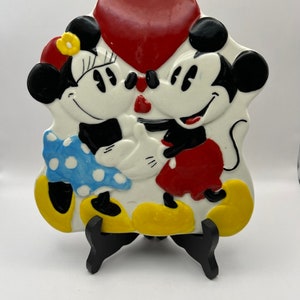 May include: A ceramic figurine of Mickey Mouse and Minnie Mouse kissing, with a red heart above them. They are both wearing their signature red and yellow outfits. The figurine is on a black stand.