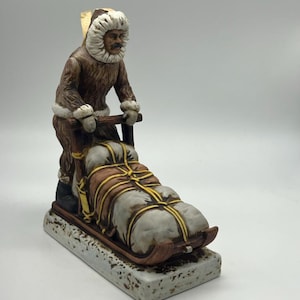 Puede incluir: Una figurita de cerámica de una persona que lleva una parka con capucha forrada de piel y sostiene un trineo con mercancías atadas. La figurita está pintada en tonos marrones, blancos y dorados.