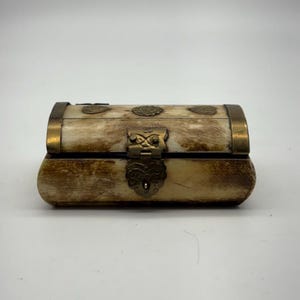 May include: A small, rectangular box with a curved top. The box is made of a light-coloured material with a mottled pattern and brass-coloured metal trim and clasp. Decorative circular accents are on the top. The box is closed.