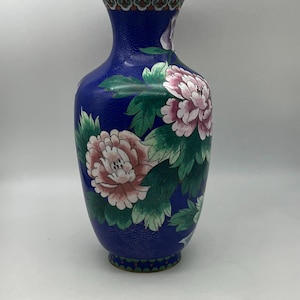 May include: A blue cloisonné vase with a gold rim and base. The vase is decorated with pink and white peony flowers and green leaves. The background has a textured pattern. The vase is approximately 25 cm tall.