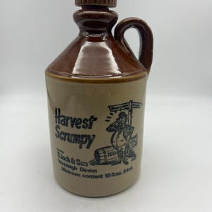 Könnte beinhalten: Eine braune Keramikkanne mit Henkel und Korkverschluss. Die Kanne hat ein Etikett mit der Aufschrift "Harvest Scrumpy" und eine Comic-Illustration eines Mannes mit einem Fass. Das Etikett enthält auch den Text "S. Linch & Son, Widdeleigh Devon, Minimum content 19 fl.oz. 54cl."