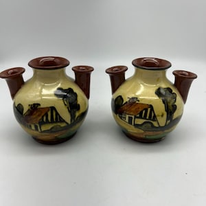 Könnte beinhalten: Zwei kleine braune Keramikvasen mit jeweils drei Ausgüssen. Die Vasen sind mit einer bemalten Szene eines Häuschens, Bäumen und eines Weges verziert.