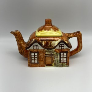 Peut inclure: Une théière en céramique brune et blanche en forme de cottage avec un toit de chaume. La théière a un petit bec verseur et une poignée et est décorée de fenêtres et d'une porte.
