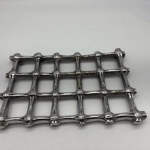 May include: A silver-coloured, rectangular decorative grate with a grid pattern. The grate is made of metal and has a series of intersecting bars forming square openings. The bars have a slightly textured surface and rounded ends.