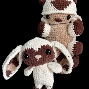May include: Two handmade crochet plush toys. One is a brown and white creature with large ears and a smiling face. The other is a similar plush toy with a hat. Both toys have black eyes and are set against a black background.