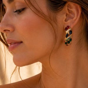 Puede incluir: Un pendiente de aro dorado con un diseño trenzado adornado con pequeñas piedras preciosas multicolores en tonos rojo, azul y verde. El pendiente cuenta con un mecanismo de cierre seguro y se lleva en el lóbulo de la oreja con acabado pulido.