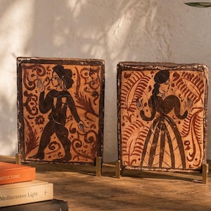Puede incluir: Dos azulejos rectangulares de terracota con figuras pintadas a mano en marrón oscuro de una persona con sombrero y otra con vestido, rodeadas de intrincados patrones de hojas. Los azulejos se exhiben en pequeños soportes de metal junto a libros.