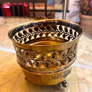 Puede incluir: Un cuenco de metal dorado vintage con un diseño decorativo de filigrana floral alrededor del borde. El cuenco tiene una base redondeada y pequeños pies. El interior del cuenco es de color dorado brillante y el exterior tiene un aspecto ligeramente envejecido.