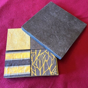 Könnte beinhalten: Ein Set aus vier quadratischen Untersetzern mit verschiedenen Designs. Die Untersetzer bestehen aus einem strukturierten Papiermaterial mit goldenen Akzenten. Die Designs umfassen ein durchgehend goldenes Quadrat, ein gestreiftes Muster, ein Baumzweigmuster und ein durchgehend schwarzes Quadrat.