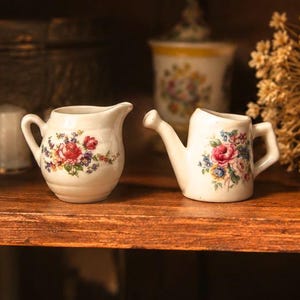 May include: Two miniature white ceramic vessels featuring colorful floral patterns with red roses and small blue and yellow flowers. One vessel is a small creamer pitcher with a curved handle, and the other is a tiny watering can with a spout.