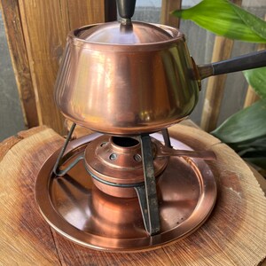 May include: A copper fondue pot with a lid and a black handle. The pot sits on a matching copper stand with a burner. The set is displayed on a wooden surface.
