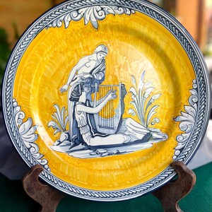 Puede incluir: Un plato grande, redondo, de cerámica amarilla con un diseño azul y blanco que representa a dos figuras tocando una lira. El plato tiene un borde azul y blanco con un patrón de cuerda.