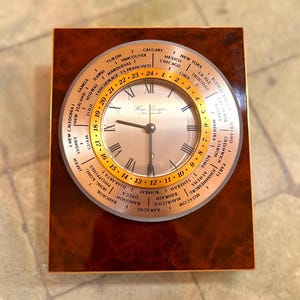 Puede incluir: Un reloj mundial de madera con un bisel giratorio dorado que muestra ciudades internacionales. La esfera del reloj tiene números romanos y la marca "Hour Lavigne". El reloj está sobre una base rectangular de madera.