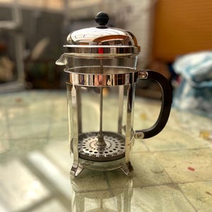 Puede incluir: Una cafetera de prensa francesa con una jarra de vidrio transparente, un marco de acero inoxidable y un asa negra. La tapa y el émbolo también son de acero inoxidable. La cafetera está sobre una superficie embaldosada.
