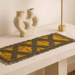 Puede incluir: Un camino de mesa rectangular de terciopelo color amarillo mostaza con un patrón entrecruzado gris oscuro y flecos a juego en los cuatro bordes. El camino de mesa está extendido, mostrando la textura de la tela y el borde decorativo.