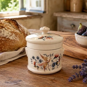Puede incluir: Un tarro de cerámica de color crema con tapa a juego, que presenta un diseño pintado de un pájaro rodeado de flores azules y naranjas. El recipiente cilíndrico tiene dos pequeñas asas laterales y se apoya sobre una superficie de madera.