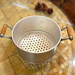 Puede incluir: Una olla vaporera vintage de aluminio con un inserto perforado. La olla tiene dos asas de madera y un acabado plateado. La vaporera está diseñada para cocinar verduras y otros alimentos. La olla está sobre una mesa.