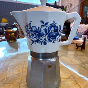 Puede incluir: Una cafetera italiana blanca y plateada. La parte superior de cerámica blanca presenta diseños florales azules. La base plateada tiene una forma geométrica. El asa y el pomo de la tapa son blancos.