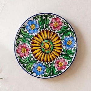 Puede incluir: Un plato decorativo redondo de cerámica con un gran motivo central de girasol amarillo rodeado de flores azules y rosas alternas. El diseño incluye intrincados patrones de follaje verde y un borde de color azul oscuro.