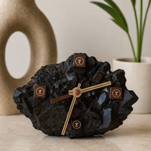 Puede incluir: Un reloj de escritorio negro, dentado y con aspecto de roca, con una superficie cristalina texturizada. Presenta cuatro marcadores cuadrados marrones con un símbolo circular en las posiciones de las doce, tres, seis y nueve, y manecillas doradas.