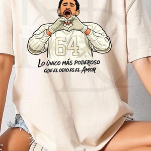 May include: Beige t-shirt featuring a cartoon illustration of a person making a heart shape with their hands. The number "64" is above the text "Lo único más poderoso que el odio es el amor."
