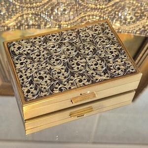 以下が含まれることがあります： 長方形の金色のコンパクト。ヒンジ付きの蓋には、黒と金の装飾的な繰り返し模様があります。コンパクトには小さな長方形の留め金が付いています。全体的なデザインはヴィンテージでエレガントです。