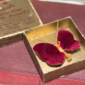 May include: A vintage velvet butterfly brooch in a gold box. The butterfly has deep red wings and a gold-coloured body. The box is open, revealing the brooch on a red surface. The box has text that reads "Coronation Velvet Butterfly".