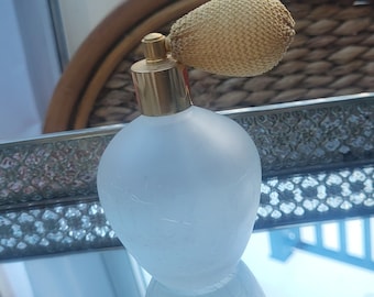 May include: A vintage perfume bottle with a frosted glass body and a gold-colored metal top. The bottle has a bulb-shaped design and a woven, beige atomizer. The bottle is sitting on a mirrored surface.