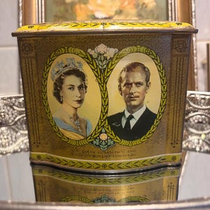 May include: A rectangular gold-toned metal tin featuring a commemorative portrait of Queen Elizabeth II and the Duke of Edinburgh within oval frames. The text below the portraits reads HM QUEEN ELIZABETH II AND HRH THE DUKE OF EDINBURGH.