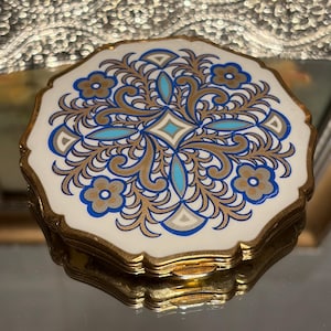 May include: A vintage compact mirror with a white, gold, and blue floral design. The compact has a scalloped edge and a gold-tone metal frame. The mirror is reflected on a mirrored surface.