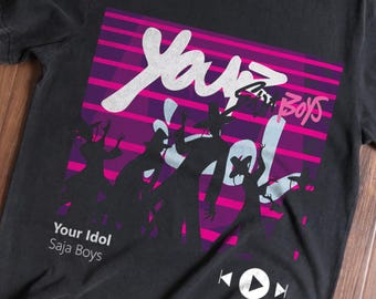 May include: Black t-shirt featuring a graphic design with the text "Your Idol" in white, above the words "Saja Boys". The design includes silhouettes of figures in a purple and pink striped background. A play button graphic is at the bottom.