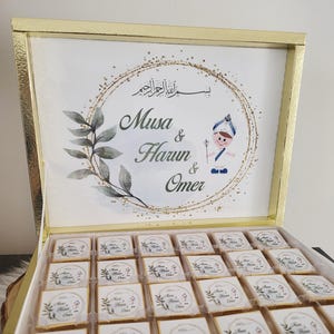 Può includere: Una scatola dorata piena di cioccolatini confezionati singolarmente. Il coperchio della scatola presenta i nomi "Musa & Harun & Omer" all'interno di un cerchio decorativo, insieme a una piccola illustrazione. I cioccolatini sono decorati con disegni simili.