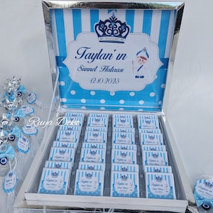 May include: A silver box with a blue and white striped lid. The lid has a crown and the text "Taylan'in Sünnet Hatırası 12.10.2023". The box is filled with small blue and white boxes with the same design as the lid.
